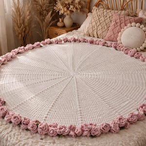 Può includere: Una coperta rotonda bianca all'uncinetto con un bordo di rose rosa. La coperta è esposta su un letto con cuscini decorativi. Il motivo all'uncinetto si irradia dal centro, creando una superficie strutturata.
