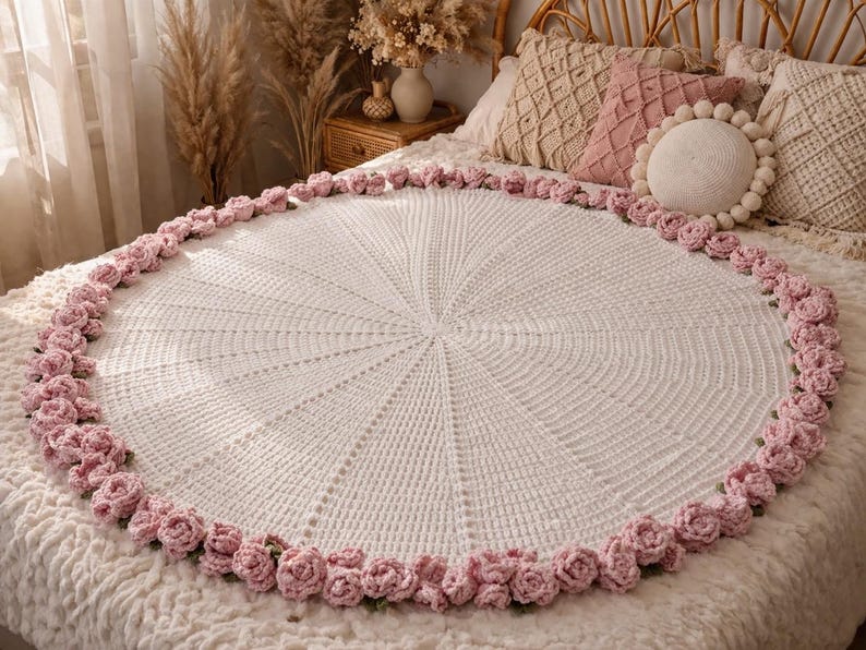 May include: A white, round, crocheted blanket with a pink rose border. The blanket is displayed on a bed with decorative pillows. The crochet pattern radiates from the center, creating a textured surface.