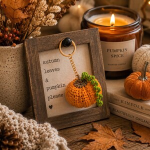 Puede incluir: Naturaleza muerta con tem&aacute;tica oto&ntilde;al con un llavero de calabaza de ganchillo, una vela de especias de calabaza encendida y calabazas decorativas. El texto enmarcado dice "autumn leaves & pumpkin please".