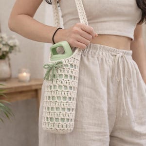 Könnte beinhalten: Ein cremefarbener, gehäkelter Wasserflaschenhalter mit passendem Riemen. Der Halter hat eine dekorative grüne Schleife und hält eine grüne Wasserflasche. Das Design ist offen, mit einem gitterartigen Muster. Der Halter ist etwa 25 cm lang.