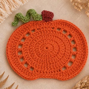 May include: An orange crochet pumpkin coaster with a green stem and a burgundy top. The coaster has a circular design with a series of small, evenly spaced holes around the edge. Perfect for fall decor.