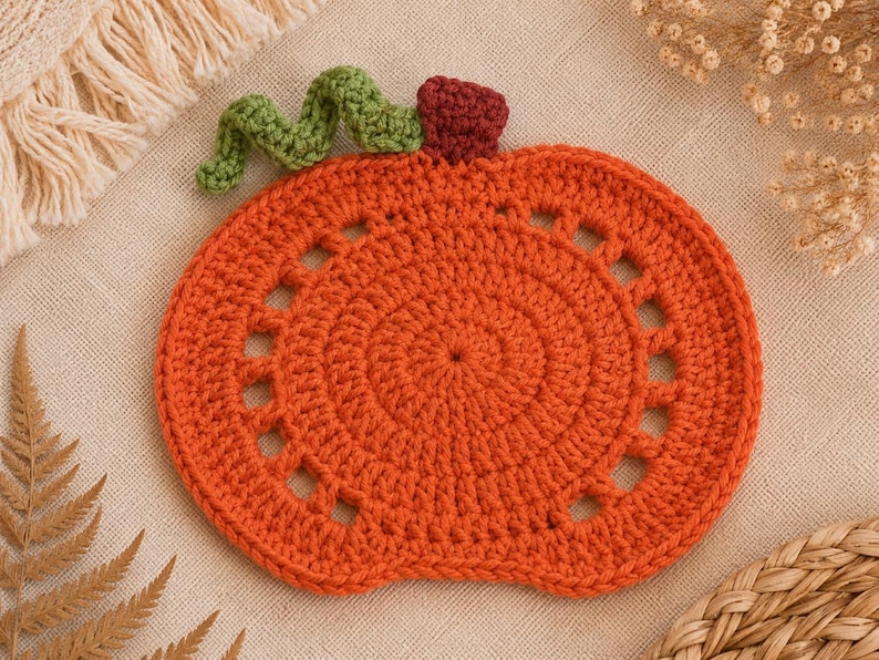 May include: An orange crochet pumpkin coaster with a green stem and a burgundy top. The coaster has a circular design with a series of small, evenly spaced holes around the edge. Perfect for fall decor.