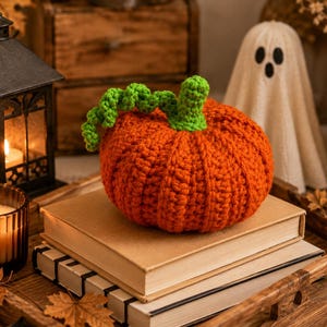 May include: A round, orange crocheted pumpkin with a textured, ribbed surface and a bright green crocheted stem featuring a curly vine. The pumpkin is crafted from yarn and sits atop a stack of two books, showcasing a handmade, soft appearance.