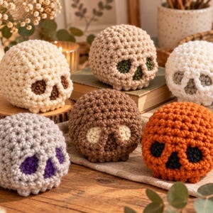 May include: A collection of six crocheted skull figures in various colors including cream, beige, white, brown, orange, and gray. Each skull features contrasting yarn colors for the eye sockets and nasal cavity, showcasing a textured, knotted stitch pattern.