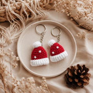 May include: Two crochet keychains shaped like miniature Santa hats. Each hat is bright red with a white brim and a white pom-pom on top. Small white snowflake-shaped embellishments are attached to the red portion of the hats. Each hat is connected to a silver-toned metal split ring by a short chain.