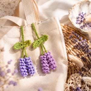 Può includere: Due rametti di lavanda all'uncinetto con foglie e steli verdi sono appoggiati su una borsa tote beige. La lavanda è di un vibrante colore viola. Lo sfondo include un cestino intrecciato, lavanda secca e un piatto decorativo.
