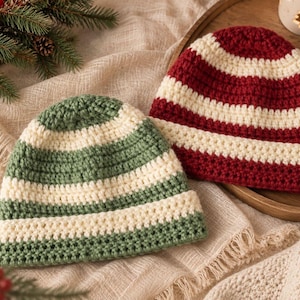 May include: Two crocheted beanies featuring horizontal stripes. One beanie has alternating bands of sage green and cream, while the other has alternating bands of deep red and cream. Both hats have a folded brim and a textured yarn construction.