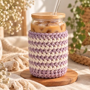 Peut inclure: Un verre transparent rempli d'une boisson glacée, doté d'une housse en crochet rayée violet clair et crème, d'un couvercle circulaire en bambou et d'une paille en verre droite. La housse présente un motif de mailles texturé sur sa partie inférieure.