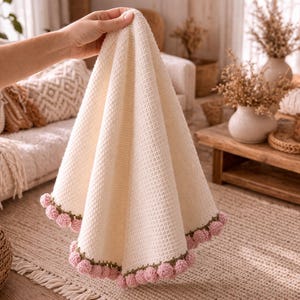 May include: Cream-colored, textured blanket with a waffle weave pattern. The blanket is trimmed with a row of pink pom-poms and green stitching. The blanket is held up by a person's hand, showcasing its soft texture and design.