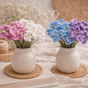 Può includere: Due vasi con fiori all'uncinetto. Uno contiene fiori rosa e bianchi, l'altro fiori blu e viola. I fiori sono in piccoli vasi di ceramica macchiettata, su sottobicchieri intrecciati. Sono visibili anche una candela accesa e una tazza di caffè.