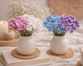 May include: Two vases with crochet flowers. One vase holds pink and white flowers, the other holds blue and purple flowers. The flowers are in small, speckled ceramic vases, sitting on woven coasters. A lit candle and a cup of coffee are also visible.