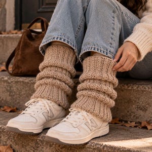 Puede incluir: Un par de calentadores de punto acanalado en color beige, usados sobre pantalones vaqueros azules y zapatillas deportivas blancas. Los calentadores presentan un patrón de punto vertical grueso y texturizado que se frunce en las piernas.