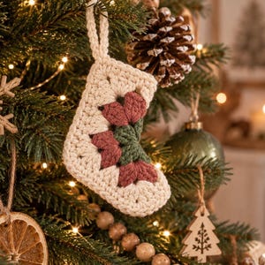 May include: A small, handmade crochet Christmas stocking ornament featuring a cream-colored yarn base with a central granny square motif in muted dusty rose and dark forest green yarn, complete with a matching cream-colored hanging loop.