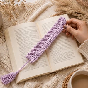 May include: A light purple crochet bookmark featuring a textured feather-like pattern and a matching tassel at the bottom. The bookmark is placed on the open pages of a book, showcasing the intricate yarn stitches and the soft lavender color.