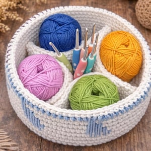 May include: A white crocheted basket with blue accents, filled with colorful yarn balls in blue, pink, green, and yellow. Crochet hooks are neatly arranged in the center. The basket is on a wooden surface.