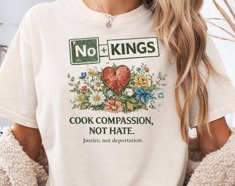 Op de afbeelding: Ivoor kleurig t-shirt met de tekst "No Kings" boven een bloemen hart ontwerp. Het shirt heeft ook de zinnen "Cook Compassion, Not Hate" en "Justice, not deportation."