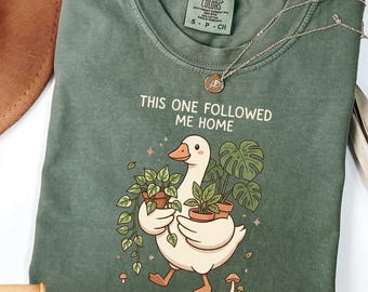 Op de afbeelding: Een mosgroen t-shirt met de tekst "THIS ONE FOLLOWED ME HOME" en een afbeelding van een witte gans met potplanten. Het shirt heeft een ronde hals en korte mouwen. Een gouden ketting ligt over het shirt.
