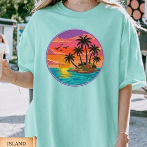 May include: A light teal t-shirt featuring a circular graphic of a tropical island scene. The design includes palm trees, a sunset, and birds. The text "ISLAND REEF" is displayed on a scroll at the bottom of the image.