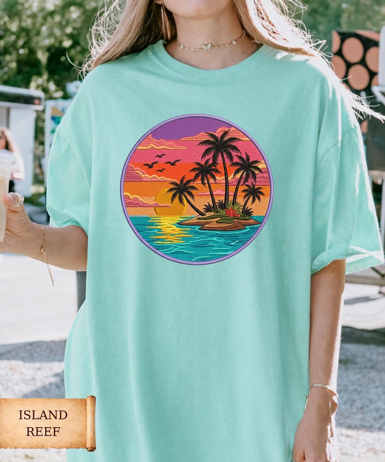 May include: A light teal t-shirt featuring a circular graphic of a tropical island scene. The design includes palm trees, a sunset, and birds. The text "ISLAND REEF" is displayed on a scroll at the bottom of the image.