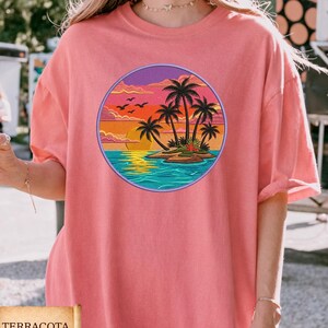 May include: Terracotta-colored t-shirt featuring a circular graphic of a tropical island scene. The design includes palm trees, a sunset, and ocean waves. The shirt has short sleeves and a relaxed fit.