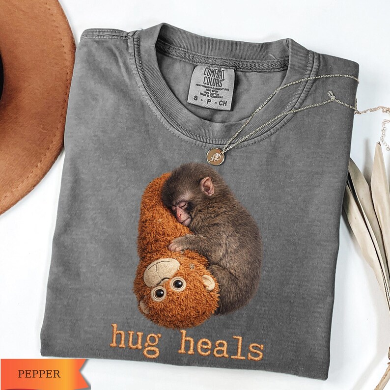 Op de afbeelding: Een grijs Comfort Colors T-shirt met een afbeelding van een slapende aap die een knuffelbeest omhelst. De tekst "hug heals" is geborduurd met oranje draad. Een zilveren ketting met een kleine hanger is over het shirt gedrapeerd.