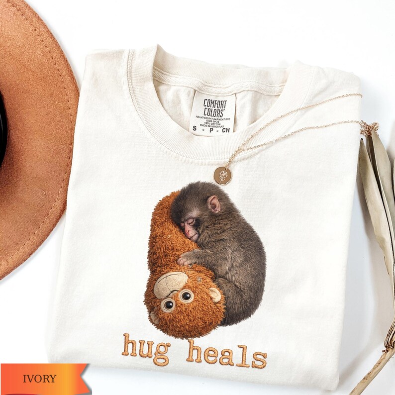 Op de afbeelding: Ivoor kleurig t-shirt met een afbeelding van een aap die een knuffelbeest omhelst. De tekst "hug heals" is geborduurd in bruin garen. Een gouden ketting met een hanger ligt op het shirt. Het label luidt "Comfort Colors".
