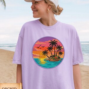 May include: A light purple t-shirt with a round graphic of a tropical island scene. The design features palm trees, a sunset, and birds. The word "ORCHID" is printed on a banner at the bottom of the image.