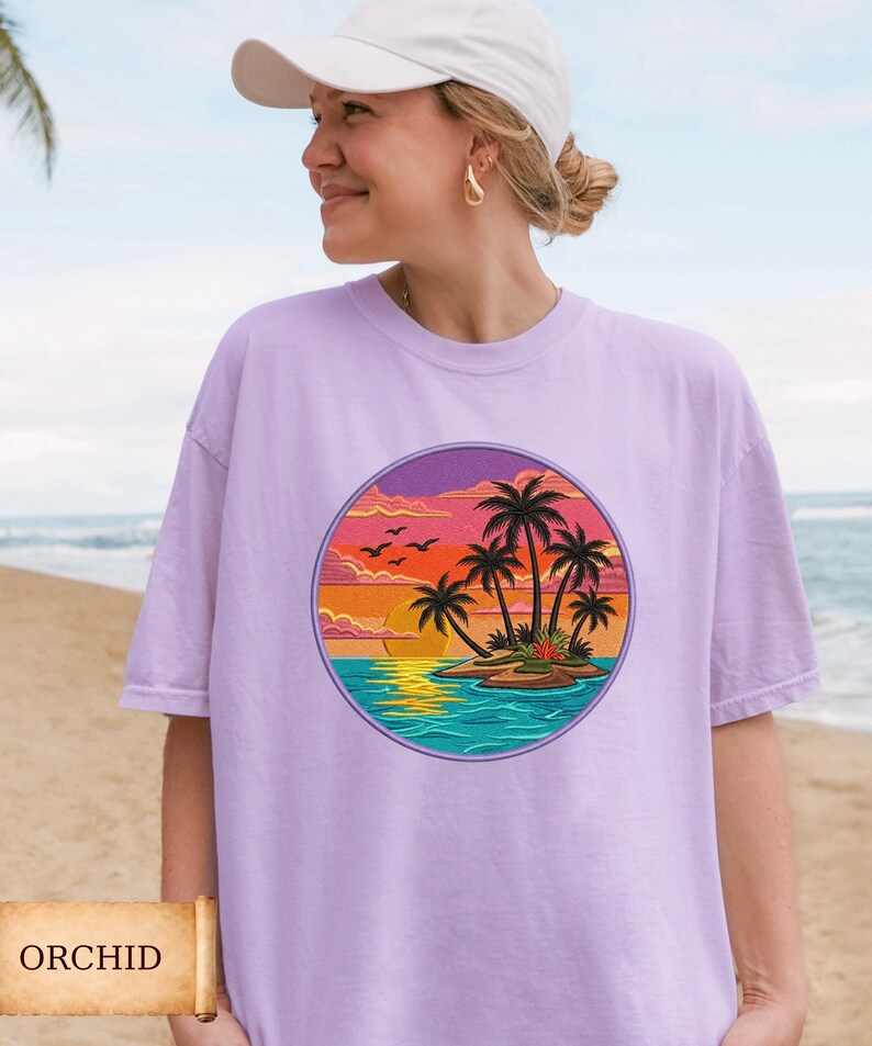 May include: A light purple t-shirt with a round graphic of a tropical island scene. The design features palm trees, a sunset, and birds. The word "ORCHID" is printed on a banner at the bottom of the image.