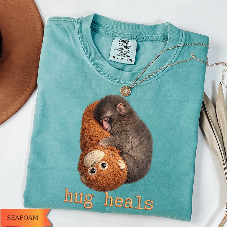 Op de afbeelding: Zeegroen t-shirt met een afbeelding van een slapende aap die een knuffelbeest omhelst. De tekst "hug heals" is geborduurd in oranje. Een gouden ketting met een hanger ligt over het shirt.