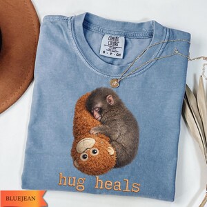Op de afbeelding: Blauw Comfort Colors T-shirt met een geborduurd beeld van een aap die een knuffel aap omhelst. De tekst "hug heals" is geborduurd met oranje draad. Een gouden ketting met een hanger is over het shirt gedrapeerd.