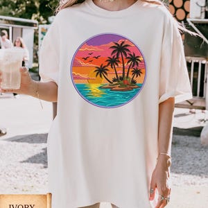 May include: Ivory-colored t-shirt featuring a circular graphic of a tropical island scene. The design includes palm trees, a sunset, and ocean waves. The shirt is a relaxed fit.