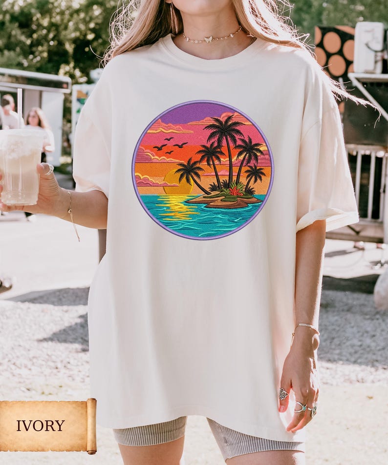 May include: Ivory-colored t-shirt featuring a circular graphic of a tropical island scene. The design includes palm trees, a sunset, and ocean waves. The shirt is a relaxed fit.