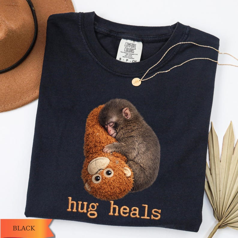 Op de afbeelding: Zwart T-shirt met een geborduurd beeld van een slapende aap die een knuffelbeest omhelst. De tekst "hug heals" is eronder geborduurd. Een gouden ketting en een bruine hoed zijn ook zichtbaar.