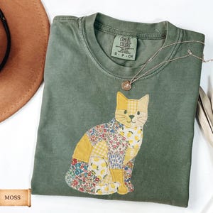 May include: A moss green t-shirt featuring a patchwork cat design. The cat is made of yellow and floral patterned fabric. A silver necklace with a pendant is on the shirt. The shirt has a Comfort Colors tag.