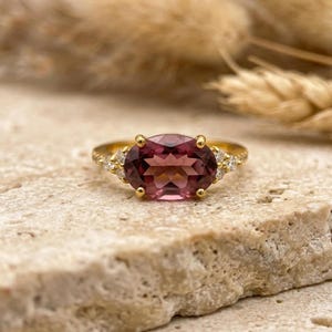 Peut inclure: Une bague en or ornée d'une grosse pierre précieuse rose de forme ovale. La bague est ornée de petites pierres précieuses claires de chaque côté de la pierre principale. La bague est posée sur une surface texturée et claire.