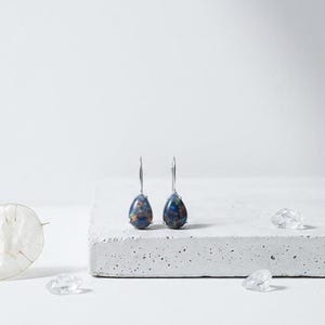 May include: A pair of teardrop-shaped earrings with a silver hook. The earrings have a dark blue and brown speckled pattern. The earrings are displayed on a white concrete surface with a white background.
