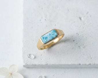 May include: A gold-colored ring with a rectangular, light blue gemstone. The ring is set on a white surface, with a flower and small, clear crystals nearby. The gemstone has a marbled appearance.
