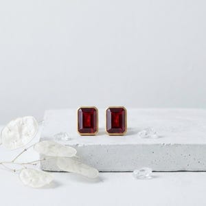 Pode incluir: Um par de brincos com pedras retangulares de cor vermelho-profundo facetadas, engastadas em armações de metal dourado. Os brincos estão posicionados lado a lado, exibindo um design geométrico limpo com um acabamento polido e contraste.