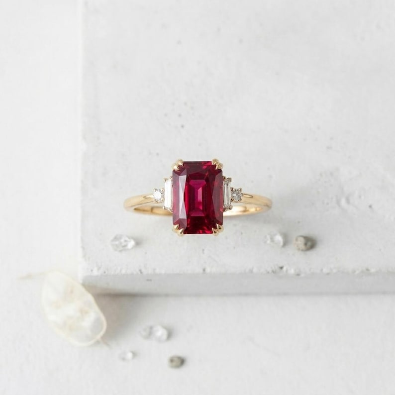 May include: A gold ring featuring a large, rectangular, emerald-cut deep red gemstone at the center. The center stone is flanked by two smaller, clear, baguette-cut stones on each side, set in a polished yellow gold band with a thin, smooth finish.