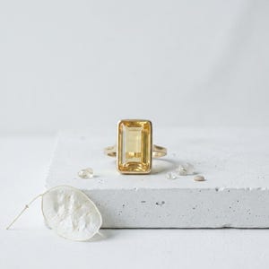 Könnte beinhalten: Ein goldener Ring mit einem großen, rechteckigen, gelben Edelstein. Der Ring liegt auf einer weißen Oberfläche, mit kleinen, klaren Kristallen daneben. Das Band des Rings ist aus poliertem Gold.