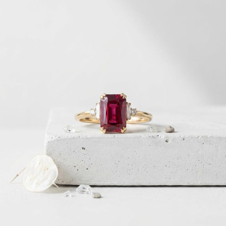 May include: A gold ring featuring a large, rectangular, emerald-cut deep red gemstone at the center, flanked by two smaller baguette-cut clear stones and two tiny round accent stones on a polished gold band.