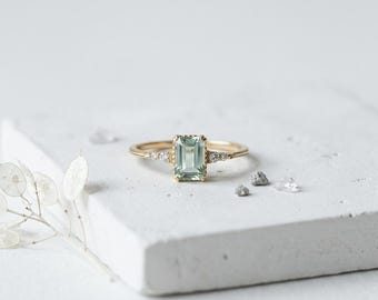 May include: A gold ring with a rectangular, light green gemstone at the center. Small, clear stones are set on either side of the main stone. The ring is displayed on a white surface, showcasing its elegant design.