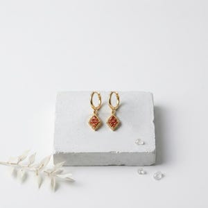 May include: A pair of gold hoop earrings featuring a dangling diamond-shaped charm. Each charm is bordered by a twisted rope texture and contains a cluster of small, vibrant orange stones set within the center of the gold frame.