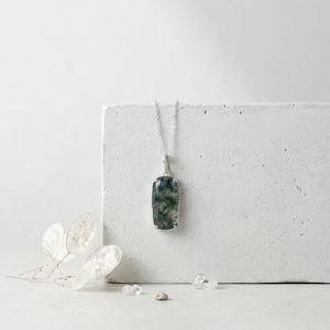 May include: A silver necklace featuring a rectangular pendant with a moss agate stone. The stone displays deep green, fern-like inclusions within a translucent, cloudy base. The pendant is held by a delicate silver chain and secured in a four-prong setting.