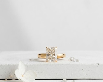 May include: A gold engagement ring with a large, rectangular-cut diamond. The ring is set on a white surface with a few small, clear crystals and a white flower. The diamond reflects light, creating a sparkling effect.
