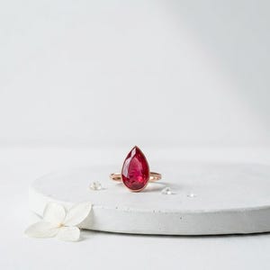 May include: A rose gold ring featuring a large, teardrop-shaped, deep red gemstone. The ring is set on a white surface with a white flower and small, clear crystals. The ring's design is simple, highlighting the vibrant color of the gemstone.