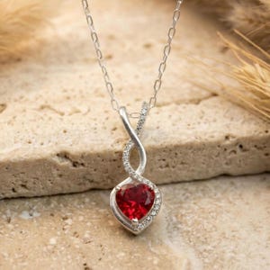 Può includere: Collana color argento con un ciondolo a forma di cuore in pietra rossa. Il ciondolo è circondato da piccole pietre trasparenti ed è sospeso a una catena delicata. Il design presenta una torsione a forma di simbolo dell'infinito sopra il cuore.