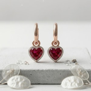 Può includere: Un paio di orecchini a cerchio in oro rosa con pendenti a forma di cuore in pietra rossa. Ogni cuore è incorniciato da un bordo delicato di piccole pietre bianche, sospeso a un cerchio in oro rosa lucido con chiusura a cerniera.