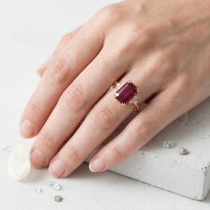 May include: A gold ring featuring a large, rectangular, deep red gemstone set in the center, flanked by two smaller, clear, baguette-cut stones on each side. The band is thin and metallic, showcasing a classic design with a prominent colored stone.