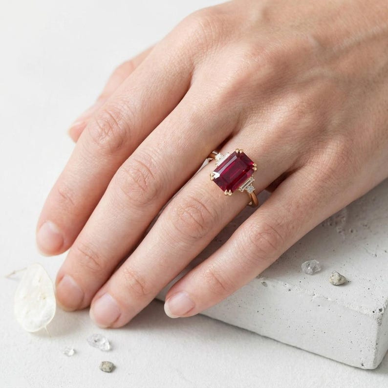 May include: A gold ring featuring a large, rectangular, deep red gemstone set in the center, flanked by two smaller, clear, baguette-cut stones on each side. The band is thin and metallic, showcasing a classic design with a prominent colored stone.